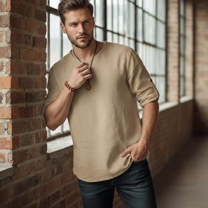 May include: A beige linen shirt with rolled-up sleeves and a round neckline. The shirt is paired with dark blue jeans. A wooden beaded bracelet and a pendant necklace complete the look. The model is standing near a brick wall.