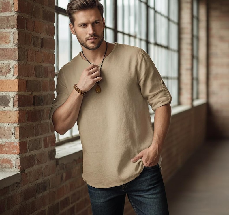 May include: A beige linen shirt with rolled-up sleeves and a round neckline. The shirt is paired with dark blue jeans. A wooden beaded bracelet and a pendant necklace complete the look. The model is standing near a brick wall.