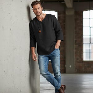 May include: Black long-sleeve shirt with a V-neck and three-quarter sleeves. The shirt is paired with light blue jeans. A pendant hangs from a black cord around the neck. The person is leaning against a concrete wall.