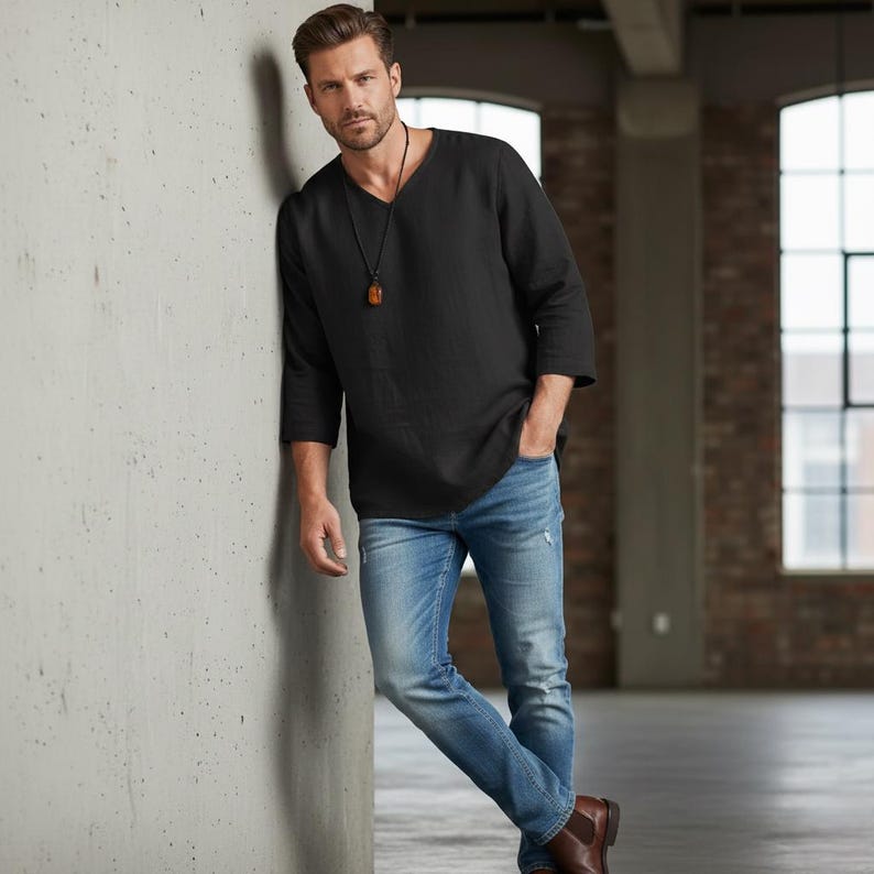 May include: Black long-sleeve shirt with a V-neck and three-quarter sleeves. The shirt is paired with light blue jeans. A pendant hangs from a black cord around the neck. The person is leaning against a concrete wall.