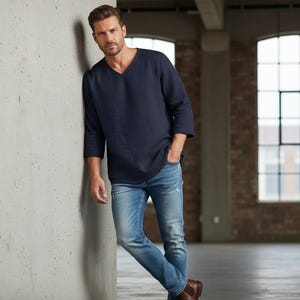 May include: A navy blue V-neck shirt with three-quarter sleeves and light blue denim jeans. The jeans have distressed details. The person is wearing brown leather boots. The background is a brick wall and a concrete wall.
