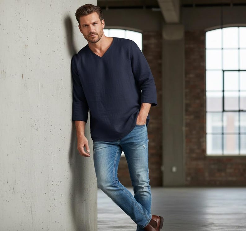 May include: A navy blue V-neck shirt with three-quarter sleeves and light blue denim jeans. The jeans have distressed details. The person is wearing brown leather boots. The background is a brick wall and a concrete wall.