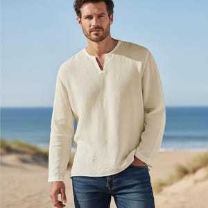 May include: A cream-coloured long-sleeved shirt with a V-neck collar. The shirt is made of a textured fabric and is paired with blue jeans. The background features a beach and ocean scene.