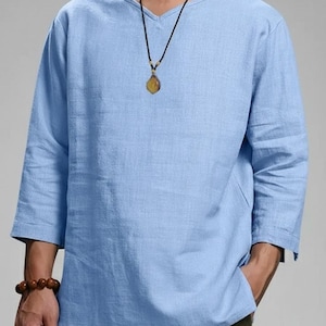 May include: A light blue, long-sleeved shirt with a V-neck. The shirt has three-quarter sleeves and a relaxed fit. A necklace with a pendant is visible. The shirt is made of a lightweight fabric, suitable for warm weather.