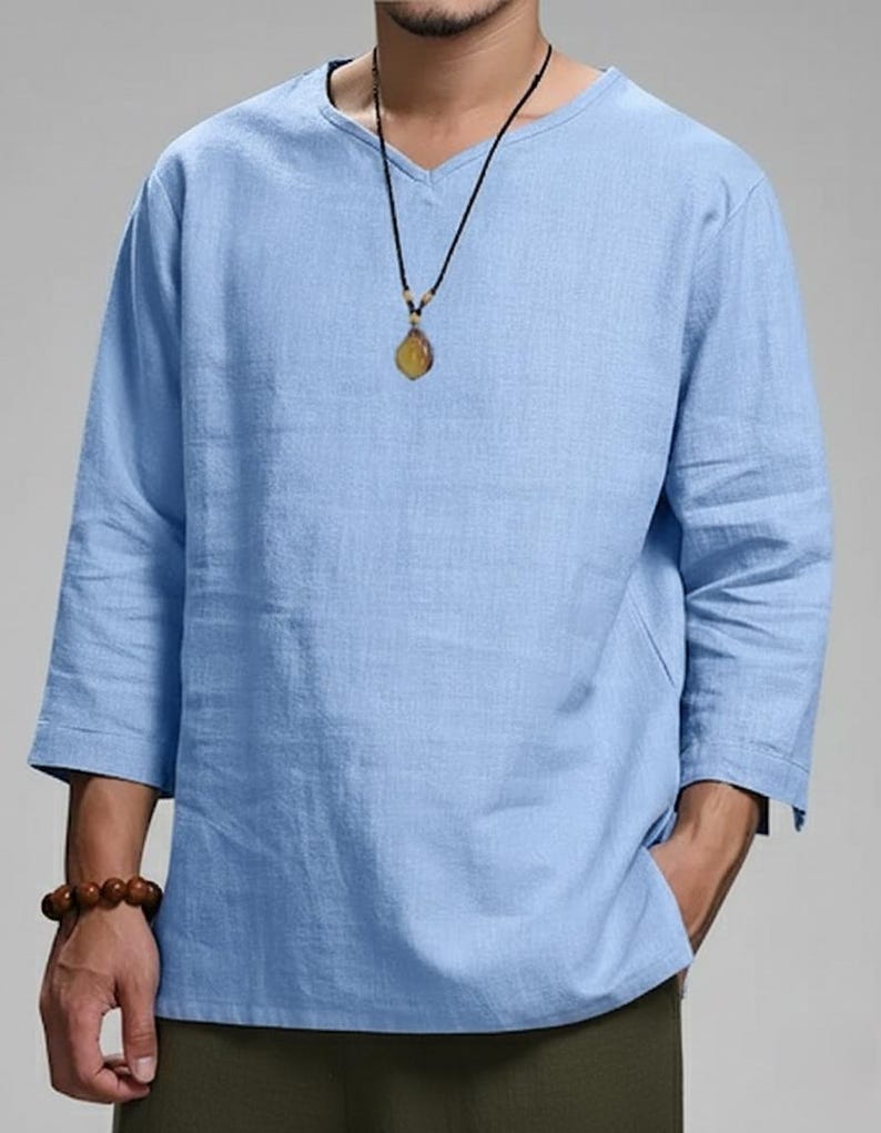May include: A light blue, long-sleeved shirt with a V-neck. The shirt has three-quarter sleeves and a relaxed fit. A necklace with a pendant is visible. The shirt is made of a lightweight fabric, suitable for warm weather.