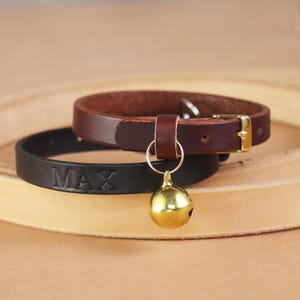 Könnte beinhalten: Ein dunkelbraunes Lederhalsband mit einer goldfarbenen Metallschnalle und einer goldfarbenen Glocke an einer Lederschlaufe, platziert vor einem schwarzen Lederhalsband mit dem eingeprägten Namen MAX in Großbuchstaben.