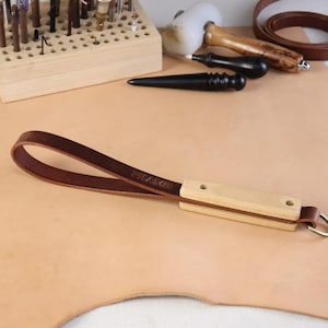 May include: A brown leather wrist strap with a light wood handle and a metal ring. The word "MIAMI" is stamped on the leather. The strap is designed for use with leatherworking tools.