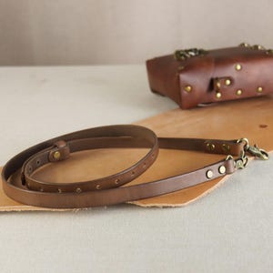 Pode incluir: Uma alça de bolsa de couro marrom com ferragens de cor latão. A alça é fina e possui uma série de pequenos orifícios ao longo do seu comprimento. Uma bolsa de couro marrom combinando é visível ao fundo.