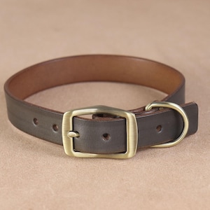 May include: A dark brown leather collar featuring a brushed brass buckle and a matching D-ring. The collar has a smooth finish with visible adjustment holes along the strap, showcasing a sturdy construction with a classic, functional design.