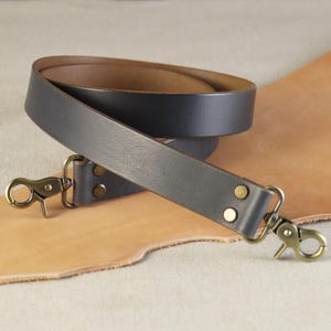 May include: A dark gray leather strap with brass-colored swivel clasps and rivets. The strap is rolled up, revealing a brown interior. The background features a tan leather surface.