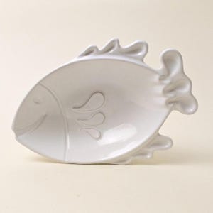 May include: A white ceramic fish-shaped dish with a smiling face and embossed details. The dish has a curved design and is suitable for serving snacks or small portions. The fish has a fin on top and a tail fin.