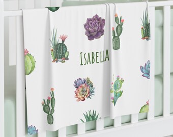 May include: White baby blanket featuring watercolor illustrations of cacti and succulents in shades of green, purple, and pink. The name "ISABELLA" is printed in green script. The blanket is draped over a white crib.