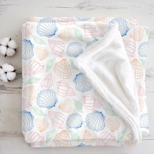 May include: A white blanket featuring a seashell pattern in blue, pink, orange, and green. The blanket has a soft, white, plush underside. The design includes various seashell illustrations.