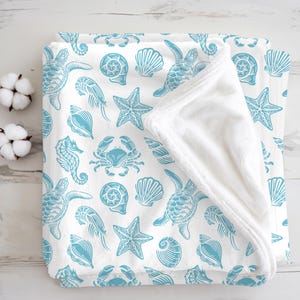 May include: A folded white blanket with a light blue sea life pattern. The pattern includes sea turtles, seashells, starfish, seahorses, and crabs. The blanket has a soft, plush texture.