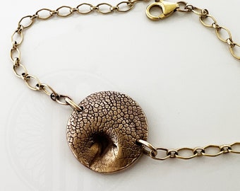 Op de afbeelding: Een gouden kettingarmband met een getextureerde ronde bedel. De bedel heeft een reliëfdesign dat lijkt op een schelp.
