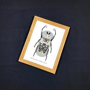 Puede incluir: Un estampado enmarcado con una ilustración estilizada de un escarabajo Goliathus Orientalis. El insecto está representado en tonos grises, negros y blancos, con un patrón detallado en su espalda. El estampado está en un marco de madera marrón claro.