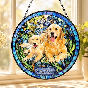 May include: A circular stained glass suncatcher featuring two golden retriever dogs resting amidst a field of blue and green iris flowers. The piece is framed in black metal with a hanging chain and includes a plaque with the text Finley & Duke.