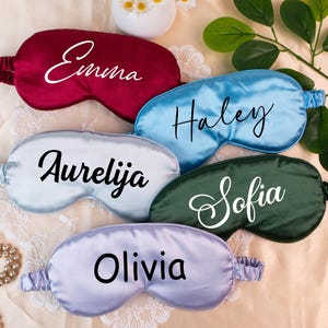 Puede incluir: Un conjunto de antifaces de satén personalizados en varios colores, como burdeos, azul, plateado, verde y lavanda. Cada antifaz presenta un nombre diferente en elegante escritura blanca: Emma, Haley, Aurelija, Sofia y Olivia. Los antifaces están sobre una superficie crema.