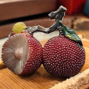 Può includere: Una scultura dettagliata di due frutti rossi, strutturati, forse litchi o simili, collegati da un gambo scuro a forma di ramo. Un frutto è tagliato, rivelando un interno dettagliato. Sono presenti anche un piccolo frutto giallo e una foglia verde.
