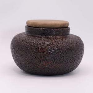 Può includere: Un vaso in ceramica marrone scuro strutturato con coperchio in legno. Il vaso ha un corpo arrotondato e un collo leggermente più stretto. Il coperchio è di forma circolare marrone chiaro, in contrasto con il corpo più scuro. Il vaso è probabilmente per la conservazione o la decorazione.