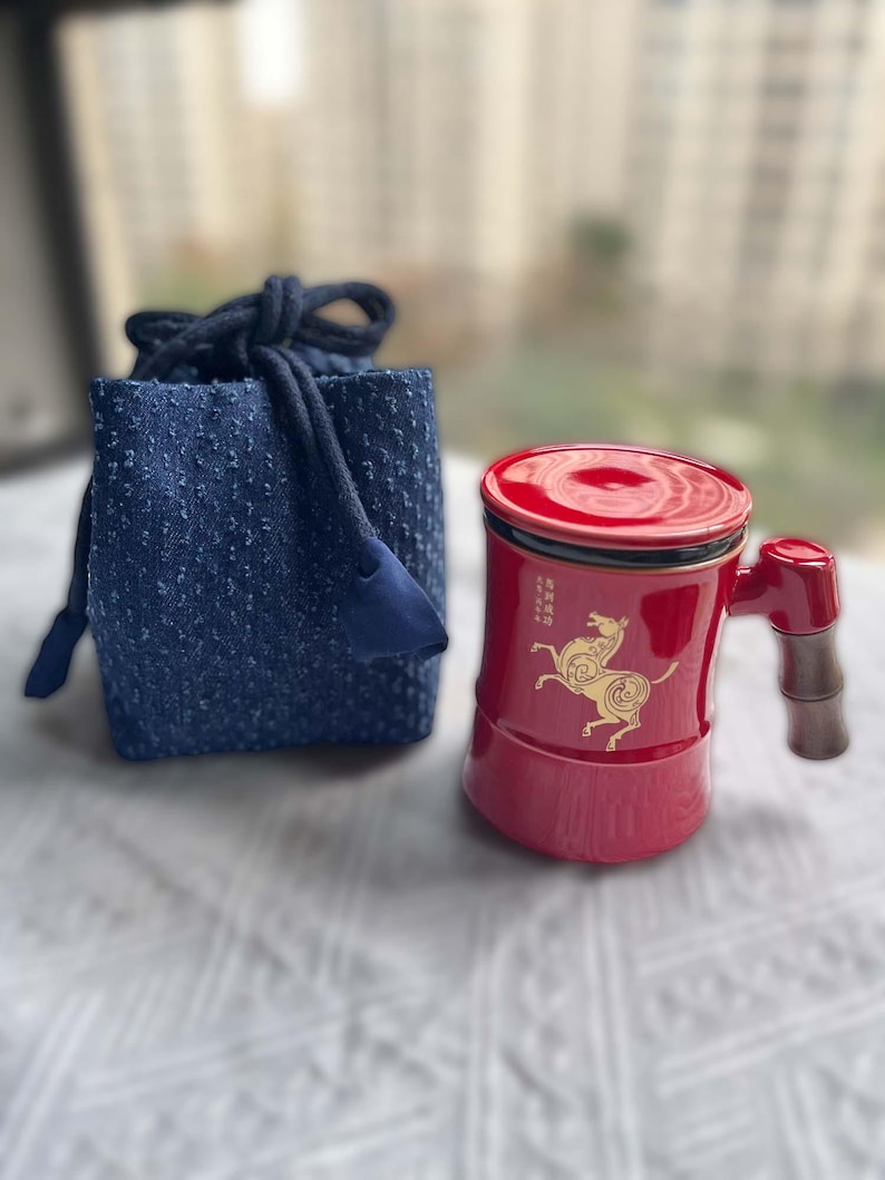 May include: A red ceramic mug with a matching lid and a bamboo-style handle, next to a small blue drawstring bag. The mug features a gold horse design and Chinese characters. The bag is made of a textured blue fabric.