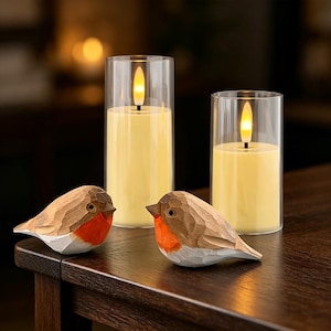 May include: Two hand-carved wooden robin figurines with orange chests and brown wings, positioned in front of two cylindrical glass candle holders containing cream-colored LED pillar candles with glowing yellow flames.