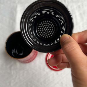 May include: A black tea infuser with a perforated interior is held above a red and black tea cup. The cup has a red exterior and a black interior. The lid is red with a black rim.