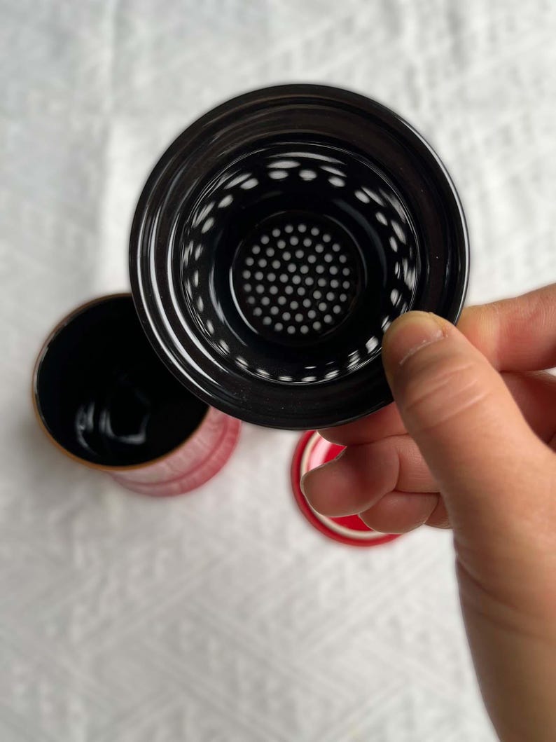 May include: A black tea infuser with a perforated interior is held above a red and black tea cup. The cup has a red exterior and a black interior. The lid is red with a black rim.