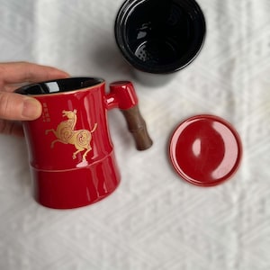 May include: A red ceramic tea mug with a black interior, a wooden handle, and a matching lid. The mug features a gold horse design and Chinese characters. A black tea infuser is also present.