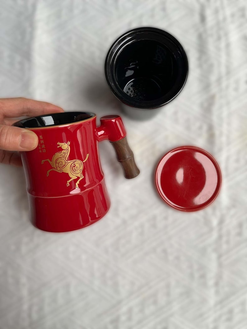 May include: A red ceramic tea mug with a black interior, a wooden handle, and a matching lid. The mug features a gold horse design and Chinese characters. A black tea infuser is also present.