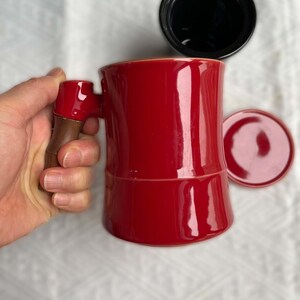 May include: A red ceramic coffee mug with a wooden handle is held by a hand. A black lid and a red saucer are next to the mug. The mug has a glossy finish and a unique shape, suitable for coffee or tea.