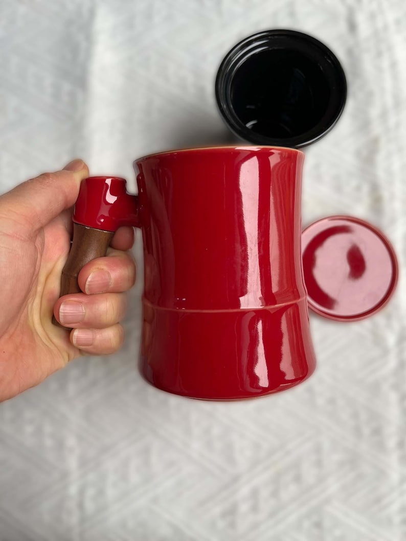 May include: A red ceramic coffee mug with a wooden handle is held by a hand. A black lid and a red saucer are next to the mug. The mug has a glossy finish and a unique shape, suitable for coffee or tea.