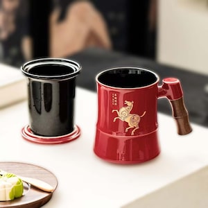 May include: A red ceramic tea mug with a gold horse graphic and a bamboo-style handle. The mug has a black interior and sits next to a black tea infuser on a red base. The mug is designed for tea lovers.