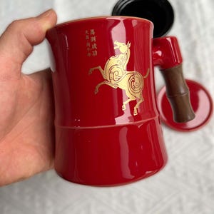May include: A red ceramic mug with a gold horse design and Chinese characters. The mug has a bamboo-style handle and a matching saucer. A black lid is also included.