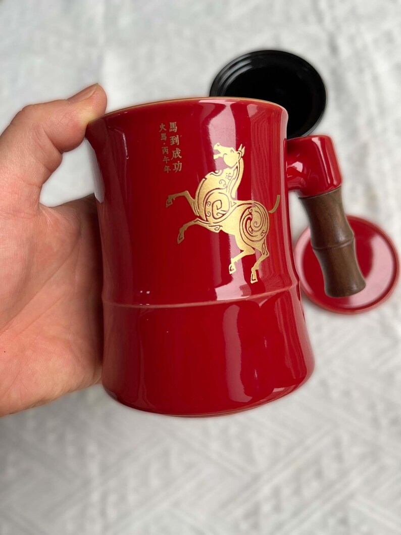 May include: A red ceramic mug with a gold horse design and Chinese characters. The mug has a bamboo-style handle and a matching saucer. A black lid is also included.