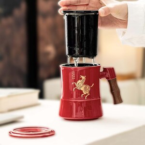 May include: A red ceramic tea mug with a gold horse design and a wooden handle. The mug has a black infuser and lid. The mug is on a white surface. The mug is approximately 4 inches tall.