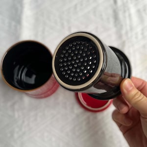 May include: Black ceramic tea infuser with a perforated lid, a red interior, and a black handle. The infuser is designed for steeping loose leaf tea. The image shows the infuser with the lid and a separate mug.
