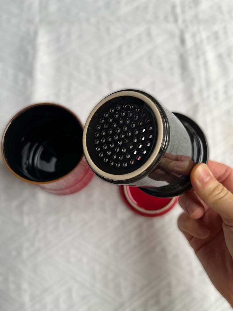 May include: Black ceramic tea infuser with a perforated lid, a red interior, and a black handle. The infuser is designed for steeping loose leaf tea. The image shows the infuser with the lid and a separate mug.