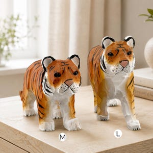Op de afbeelding: Twee handgesneden houten tijgerbeeldjes met oranje vacht met zwarte strepen, witte buiken en roze neuzen. Het kleinere beeldje is gemarkeerd met de letter M en het grotere beeldje met de letter L, wat de verschillende maten aangeeft.