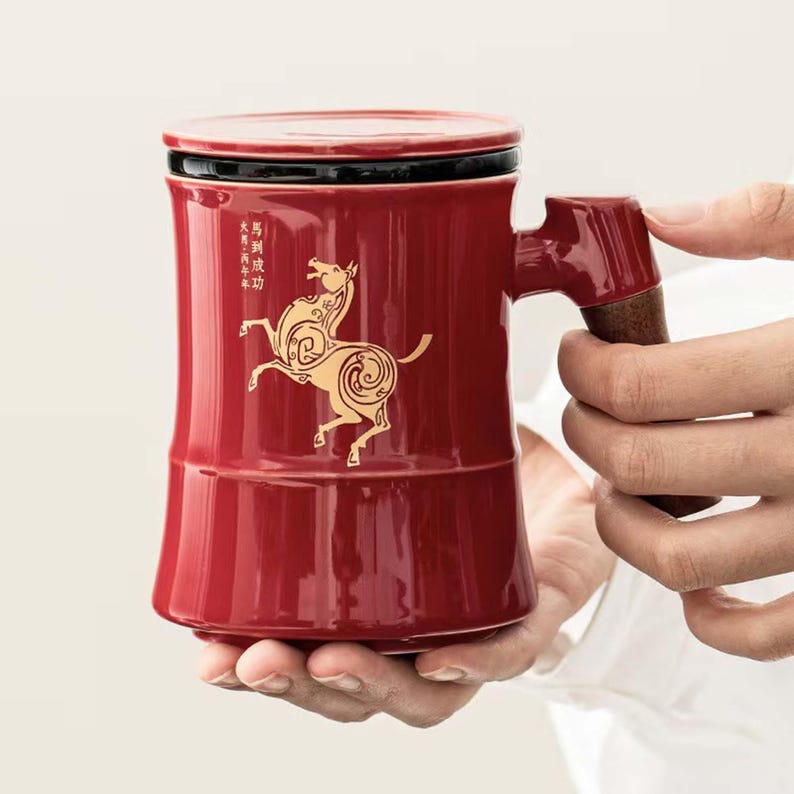 May include: A red ceramic mug with a lid and a handle. The mug features a gold horse design and Chinese characters. The handle has a wooden accent. The mug is held by a person.