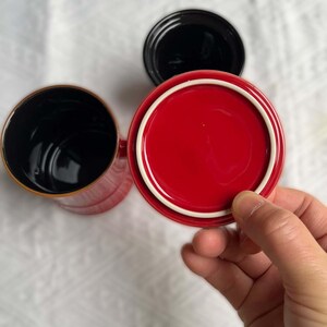 May include: Overhead shot of three ceramic mugs. One red mug with a white rim and a matching lid is held by a hand. The other two mugs are black. The mugs are on a white surface.
