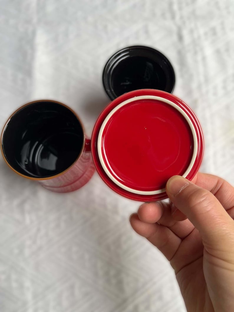 May include: Overhead shot of three ceramic mugs. One red mug with a white rim and a matching lid is held by a hand. The other two mugs are black. The mugs are on a white surface.