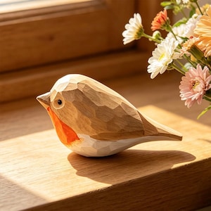 May include: A hand-carved wooden bird figurine featuring a faceted texture, a natural light brown body, a bright orange chest, and a white underbelly. The bird has a small dark eye and a pointed beak, showcasing visible chisel marks throughout.