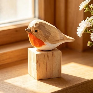 May include: A hand-carved wooden robin figurine featuring a light brown body with a distinct orange-red chest and a dark eye. The bird is perched atop a solid, light-coloured wooden cube base, showcasing visible carved facets and natural wood grain.