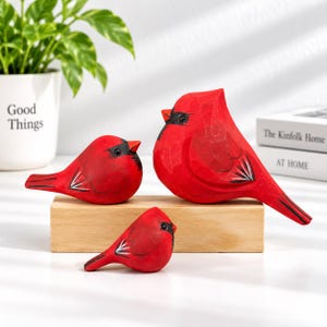 Op de afbeelding: Drie handgesneden houten vogelbeeldjes, geschilderd in felrood met zwarte gezichtsmarkeringen en vleugeldetails. De vogels zijn geplaatst op een rechthoekig lichtgekleurd houten blok, met twee vogels bovenop en één kleinere vogel aan de voorzijde.