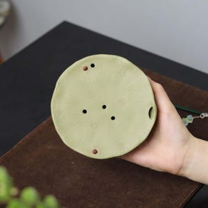Puede incluir: Una bandeja de té de cerámica verde claro con forma circular. La bandeja tiene varios agujeros pequeños y patrones decorativos. Cuenta con un pequeño asa y se sostiene en una mano.