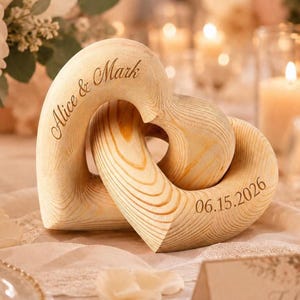 May include: Two interlocking light-colored wooden hearts with a visible natural grain pattern. The left heart is engraved with the text Alice & Mark, and the right heart is engraved with the date 06.15.2026 in a dark, elegant script font.