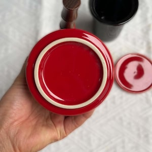 May include: A red ceramic mug with a white rim and a wooden handle. A black ceramic container and a small red lid are also visible. The items are on a white surface.
