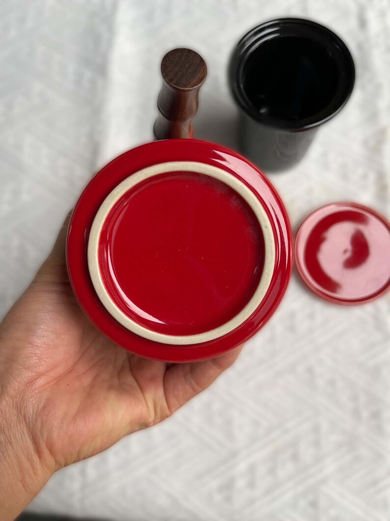 May include: A red ceramic mug with a white rim and a wooden handle. A black ceramic container and a small red lid are also visible. The items are on a white surface.