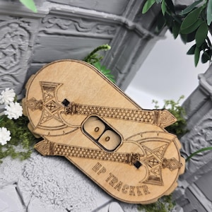 Könnte beinhalten: Ein HP-Tracker aus hellem Holz mit zwei lasergravierten, gekreuzten Streitäxten. Das Gerät verfügt über einen zentralen rechteckigen Ausschnitt mit zwei kleinen Drehscheiben, die die Zahl Null anzeigen, sowie die Gravur HP TRACKER.
